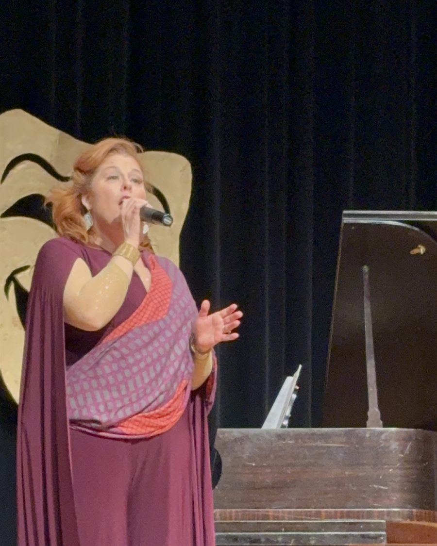 Jenn Quader singing into a microphone beside a grand piano onstage at North Lenoir's Benefit Concert, one hand open in front of her, wearing a deep purple gown with a patterned sash.