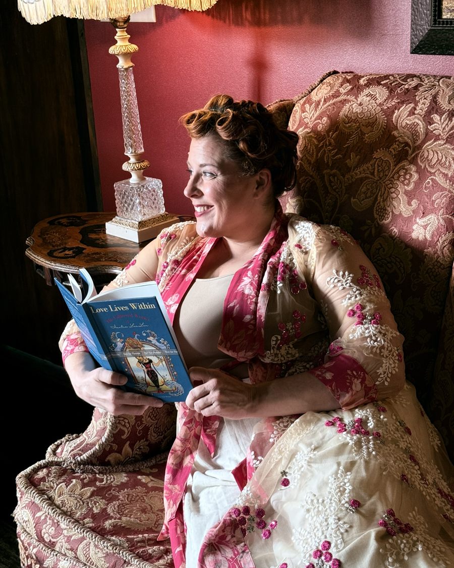 Jenn Quader smiling in a rose-patterned wingback chair beside a crystal lamp, holding the blue hardcover of Love Lives Within open on her lap, its illustrated cover facing the camera.