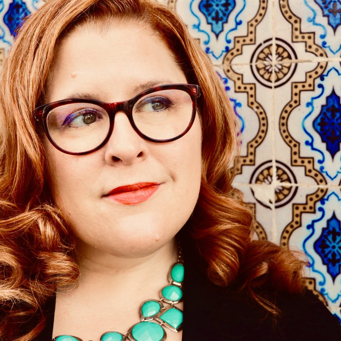 Jenn Quader in tortoiseshell glasses and a turquoise necklace, looking off to one side in front of a wall of blue and white Portuguese tiles.