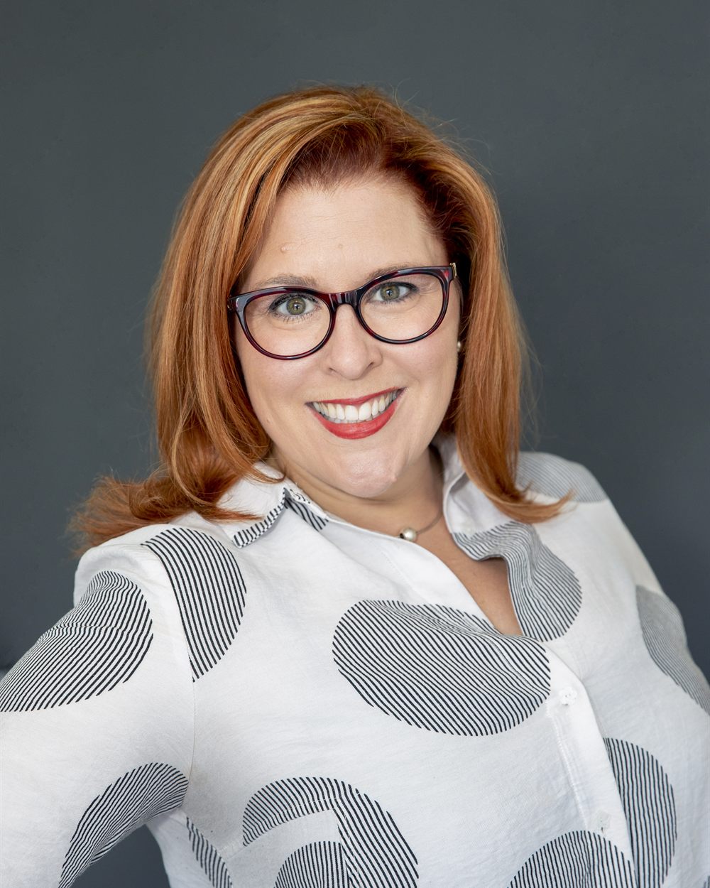 Jenn Quader in a blue patterned blouse, standing in front of a bookshelf.