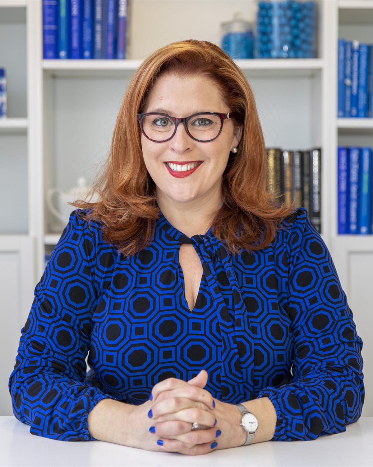 Jenn Quader seated at a white table in a patterned cobalt blouse, hands folded, smiling directly at the camera in front of a shelf of blue books.