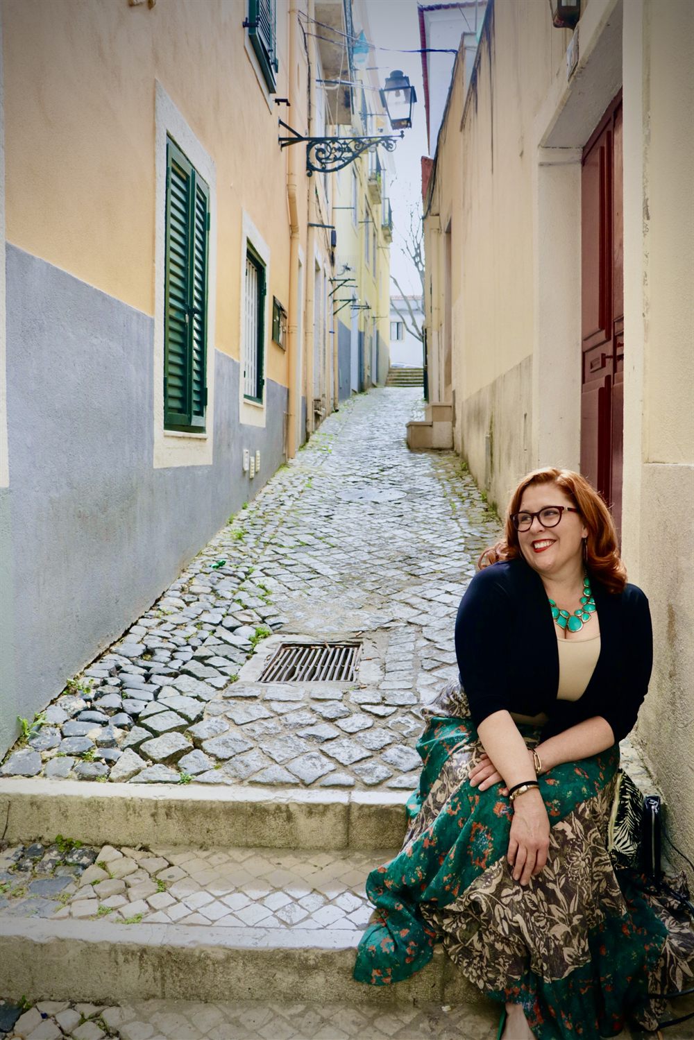 Jenn Quader sitting on stone steps in a narrow Lisbon lane, chin resting on her hand, cobblestones running away behind her.
