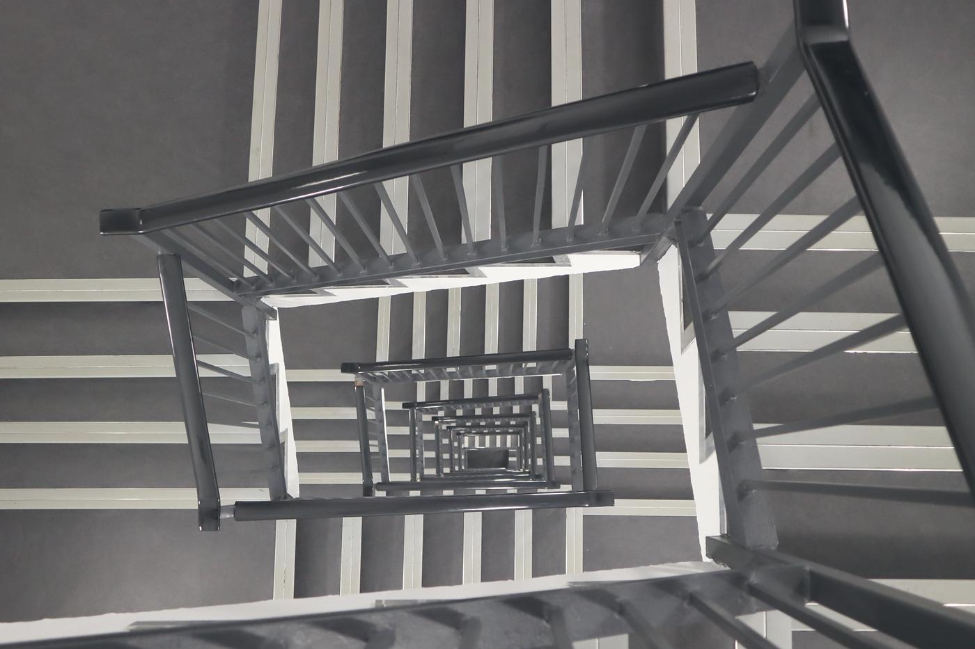 The view straight down a square stairwell, flight after flight of dark handrail shrinking away.