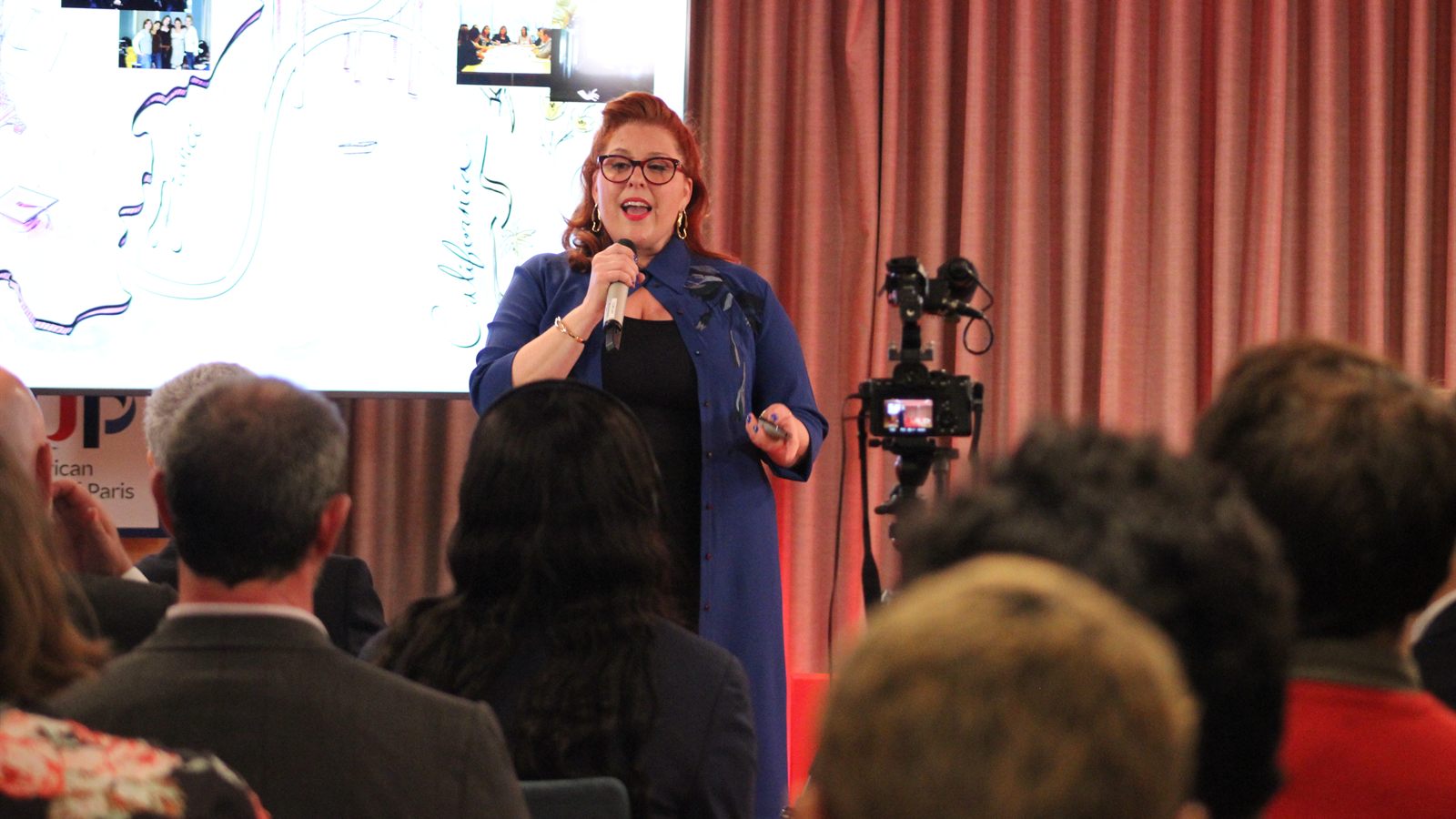 Jenn Quader speaking with a handheld microphone in front of a seated audience, her slides on the screen behind her and an American University of Paris banner to one side.