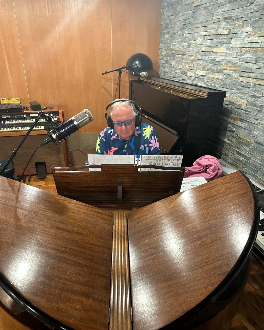 Dr. Dennis Michels at the piano in headphones and a bright patterned shirt, seen from across the open lid of a grand piano.