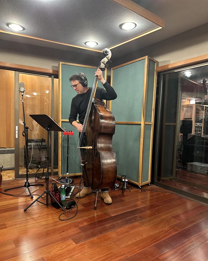 Sean Hurley standing at an upright bass in the live room, headphones on, a music stand and vocal microphone beside him.