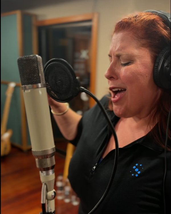 Jenn Quader singing into a large condenser microphone behind a pop filter, wearing studio headphones, eyes closed.