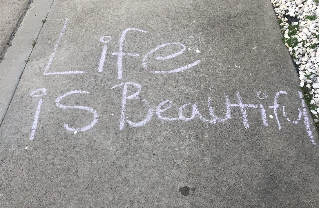 The words Life is Beautiful written in blue chalk on a sidewalk beside a border of white stones.