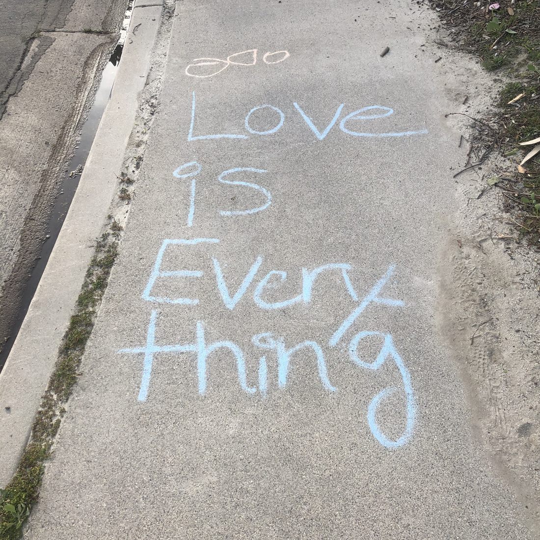 The words Love is Everything written in pale blue chalk on a concrete sidewalk.